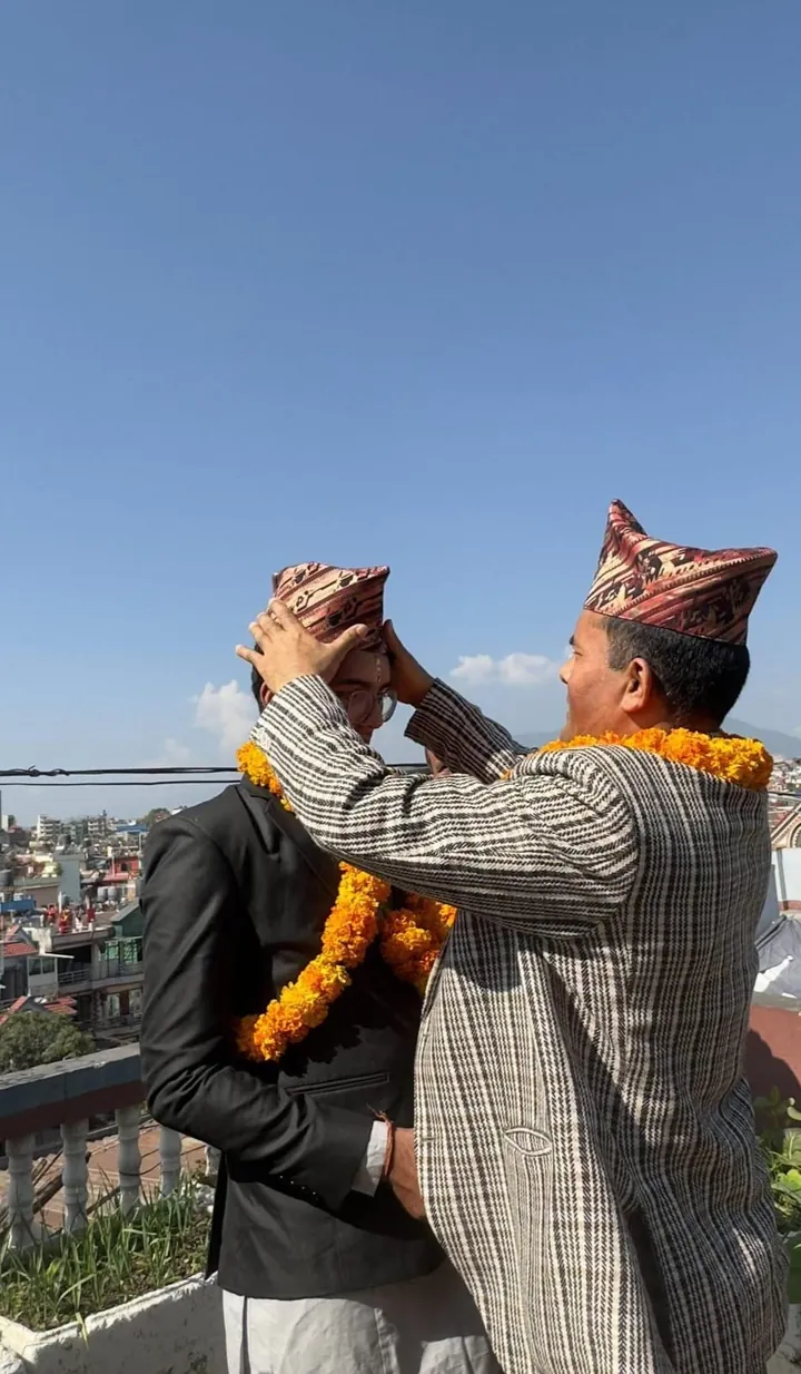 Martin's father placing a traditional topi on Martin's head during Tihar, both wearing marigold garlands